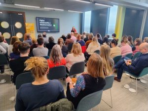 A room of people sitting down with their backs to the camera, facing towards a screen and presenter Hannah Smith